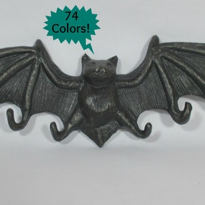 May include: A dark gray bat-shaped wall hook with outstretched wings and four hooks. The bat has detailed wing and body features. A teal speech bubble in the upper right corner says "74 Colors!"