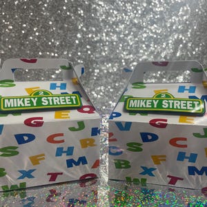 May include: Two white cardboard gable boxes featuring a colorful pattern of scattered alphabet letters in red, blue, green, and yellow. Each box displays a green and yellow rectangular sign with the text MIKEY STREET and the number 2.