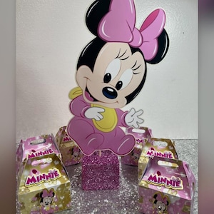 May include: A Minnie Mouse-themed party decoration set. It includes a large, pink Minnie Mouse cutout, a glittery pink base, and several small gift boxes with Minnie Mouse graphics. The image has the Instagram handle @rivera_decor_service.