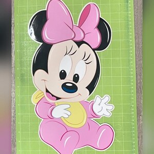 May include: A pink Minnie Mouse cutout, featuring a large bow, black ears, and a yellow bib. The character is seated on a green cutting mat. The Cricut logo is visible in the top left corner. The Instagram handle @rivera_decor_service is at the bottom.