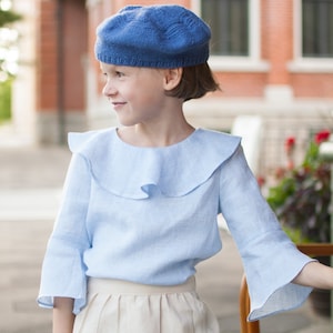 Może przedstawiać: Młoda osoba w niebieskim beretcie z dzianiny i jasnoniebieskiej lnianej bluzce z falbaną i rozkloszowanymi rękawami. Strój zawiera beżową spódnicę. Tło przedstawia ceglany budynek i miejsca do siedzenia na zewnątrz.