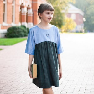 Puede incluir: Un vestido azul y verde a cuadros con mangas cortas. El vestido tiene un cuello redondo y lo lleva una persona. La persona sostiene un libro. El fondo incluye un edificio de ladrillo y una pasarela de ladrillo.