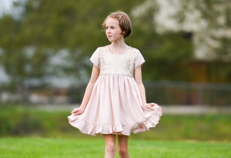May include: A light pink dress featuring a sequined bodice with short sleeves and a gathered skirt with a ruffled hemline. The dress has a round neckline and a soft, flowing silhouette that falls above the knee.