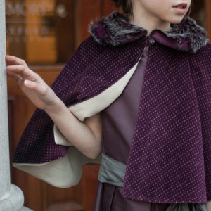 May include: A purple velvet cape with a white lining and a faux fur collar. The cape has a dotted pattern and is worn by a child.