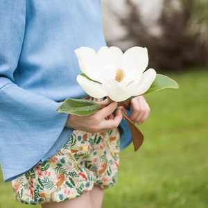 Puede incluir: Top azul claro con mangas acampanadas y shorts estampados. Los shorts tienen un estampado colorido con motivos florales y animales. Se sostiene una gran flor de magnolia blanca. Fondo verde.