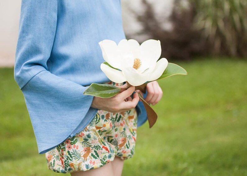 Puede incluir: Top azul claro con mangas acampanadas y shorts estampados. Los shorts tienen un estampado colorido con motivos florales y animales. Se sostiene una gran flor de magnolia blanca. Fondo verde.