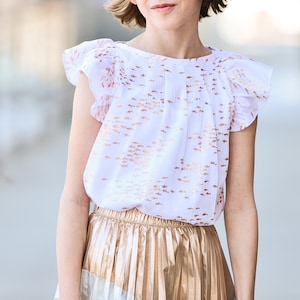 Puede incluir: Una blusa blanca con estampado de peces dorados y mangas con volantes combinada con una falda plisada dorada.