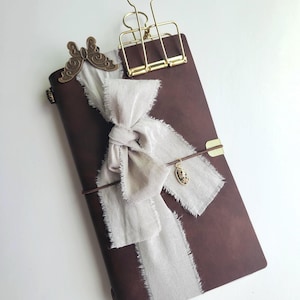 May include: A brown leather journal with a gold clip and butterfly accent. A cream-colored ribbon is tied around the journal, and a small charm hangs from a leather strap. The journal is closed.