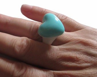 Può includere: Un anello a forma di cuore color turchese su un dito. L'anello è bianco con un cuore turchese in cima.