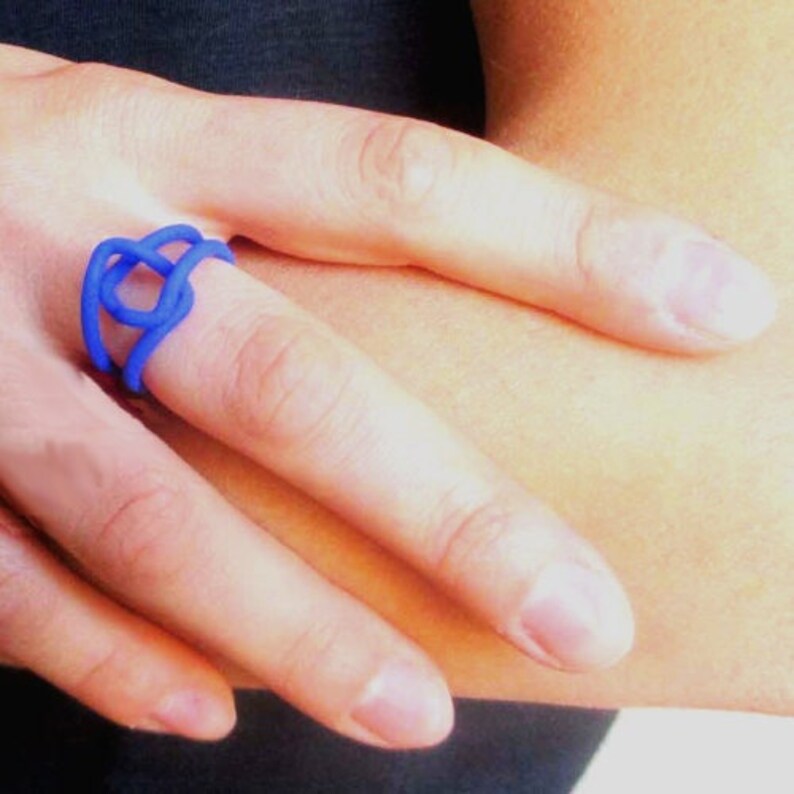 Puede incluir: Un anillo azul hecho de una sola pieza continua de metal, retorcido y anudado para crear un dise&ntilde;o &uacute;nico.