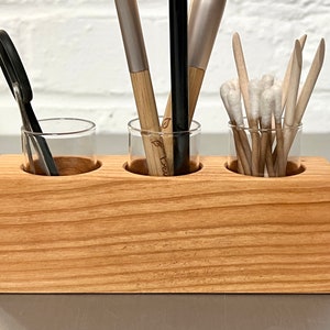 May include: A wooden organizer with three glass jars for storing makeup brushes, cotton swabs, and other small items. The organizer is made of light brown wood and has a natural finish.