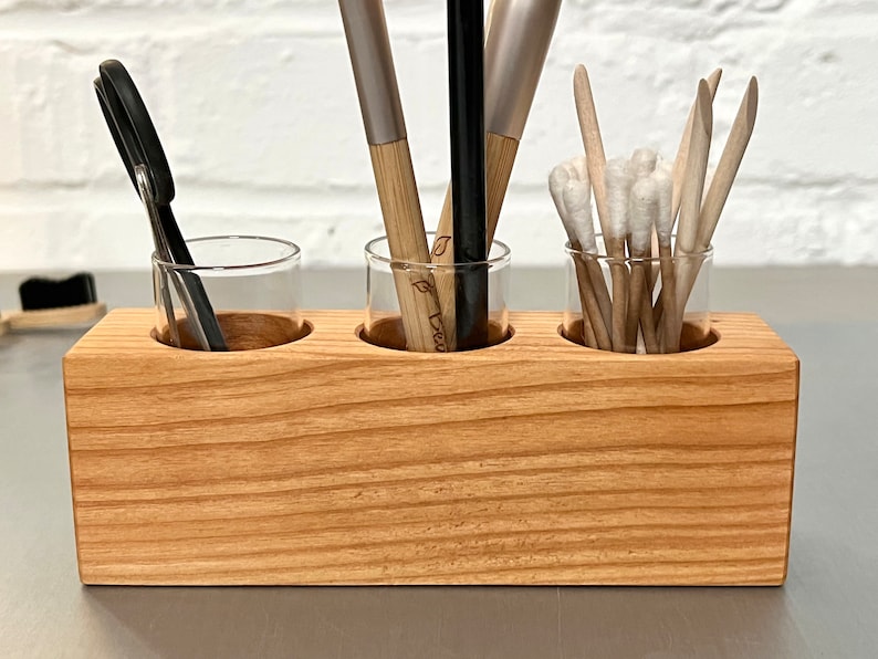 May include: A wooden organizer with three glass jars for storing makeup brushes, cotton swabs, and other small items. The organizer is made of light brown wood and has a natural finish.