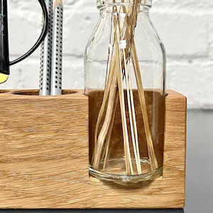 May include: A wooden desk organizer with a clear glass bottle holding dried flowers and a section for pens and a black desk lamp. The organizer is made of light brown wood.