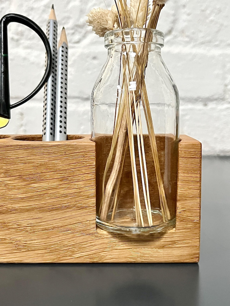May include: A wooden desk organizer with a clear glass bottle holding dried flowers and a section for pens and a black desk lamp. The organizer is made of light brown wood.