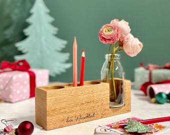 May include: A rectangular light-colored wooden block with three circular holes for pencils and a glass bottle insert holding three pink ranunculus flowers. The front of the wood features the engraved text "dein Wunschtext".
