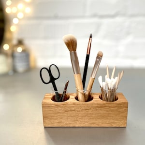 May include: Wooden makeup brush holder with three glass jars. The holder is made of light brown wood and has a natural finish. The jars are clear glass and are filled with makeup brushes, cotton swabs, and a pair of tweezers.