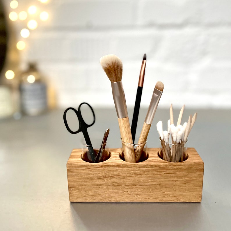 May include: Wooden makeup brush holder with three glass jars. The holder is made of light brown wood and has a natural finish. The jars are clear glass and are filled with makeup brushes, cotton swabs, and a pair of tweezers.