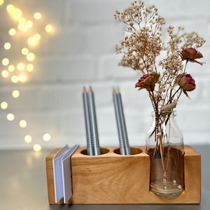 May include: A wooden desk organizer with a clear glass bottle holding dried flowers and a small stack of white cards. Two gray pencils are in the organizer. The wood has a natural finish.