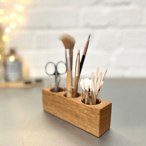 May include: Wooden organizer with three compartments for holding makeup brushes and cotton swabs. The organizer is made of light brown wood and has a natural finish.