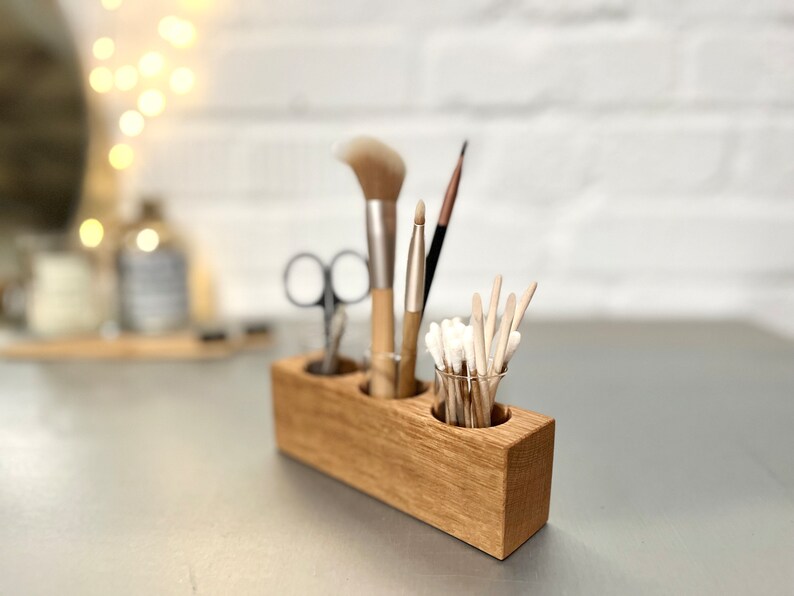 May include: Wooden organizer with three compartments for holding makeup brushes and cotton swabs. The organizer is made of light brown wood and has a natural finish.