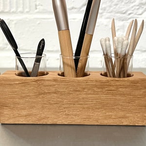 May include: A wooden organizer with three glass jars for storing makeup brushes, tweezers, and cotton swabs. The organizer is made of light brown wood and has a natural wood grain.