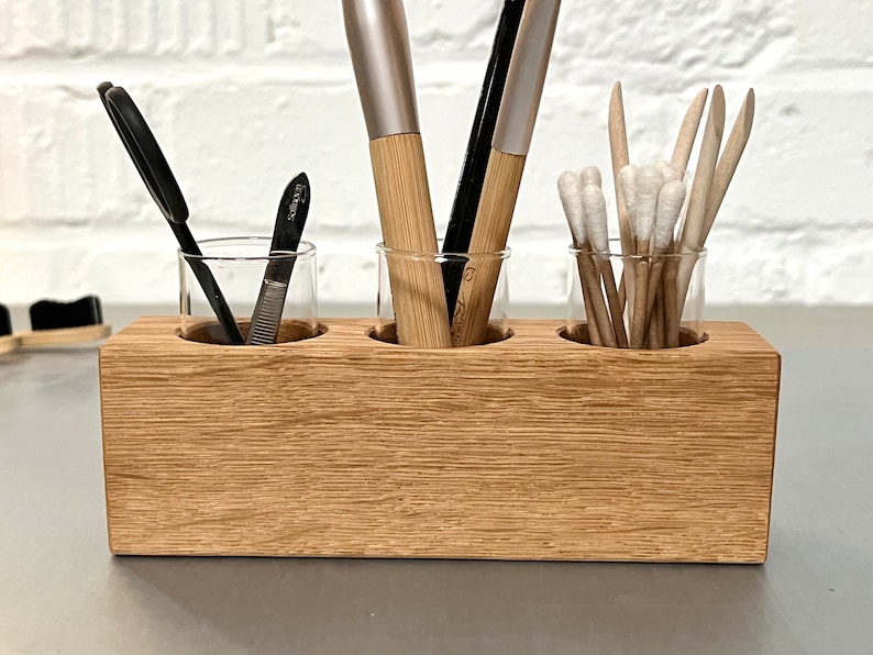 May include: A wooden organizer with three glass jars for storing makeup brushes, tweezers, and cotton swabs. The organizer is made of light brown wood and has a natural wood grain.