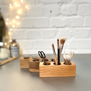 May include: Wooden makeup brush holder with five clear glass jars. The holder is made of light brown wood and has a natural finish. The jars are filled with makeup brushes, cotton swabs, and a pair of scissors.