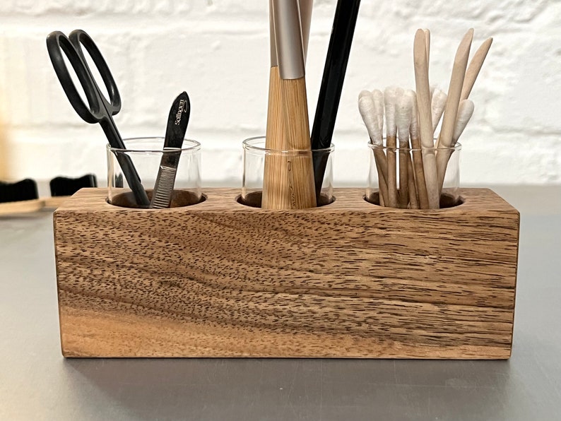 May include: A wooden organizer with three glass jars for holding small items. The jars are filled with various tools, including a pair of scissors, a nail file, and several thin wooden sticks.