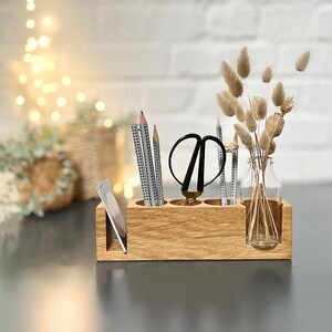 May include: A wooden desk organizer with compartments for pencils, scissors, and a small glass bottle with dried flowers. The organizer is made of light-colored wood and sits on a dark surface. The background includes a blurred view of a brick wall and decorative lights.