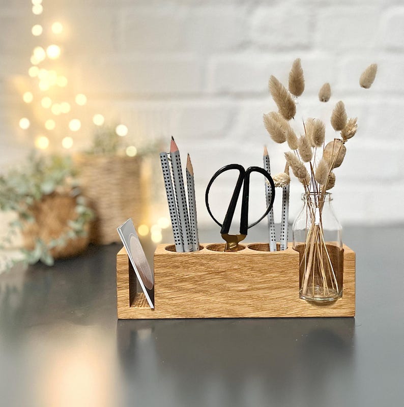 May include: A wooden desk organizer with compartments for pencils, scissors, and a small glass bottle with dried flowers. The organizer is made of light-colored wood and sits on a dark surface. The background includes a blurred view of a brick wall and decorative lights.