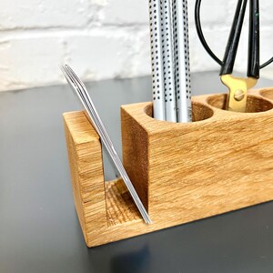May include: A wooden desk organizer with multiple compartments. It holds silver pens, a pair of scissors, and a stack of business cards. The organizer is made of light-colored wood and sits on a dark surface.