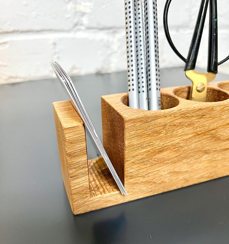 May include: A wooden desk organizer with multiple compartments. It holds silver pens, a pair of scissors, and a stack of business cards. The organizer is made of light-colored wood and sits on a dark surface.