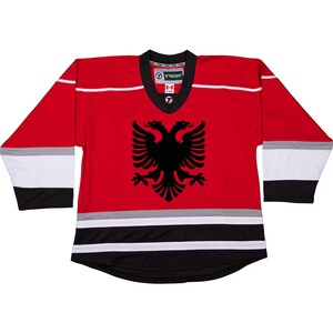 Puede incluir: Jersey de hockey rojo, blanco y negro con la bandera albanesa en la parte delantera. La bandera presenta un &aacute;guila bic&eacute;fala negra sobre un fondo rojo.