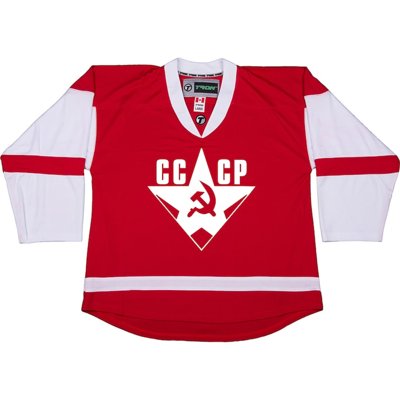 Puede incluir: Una camiseta de hockey roja con mangas blancas y un ribete blanco alrededor de la parte inferior. La camiseta tiene una gran estrella blanca con un martillo y una hoz dentro y el texto "CCCP" encima.