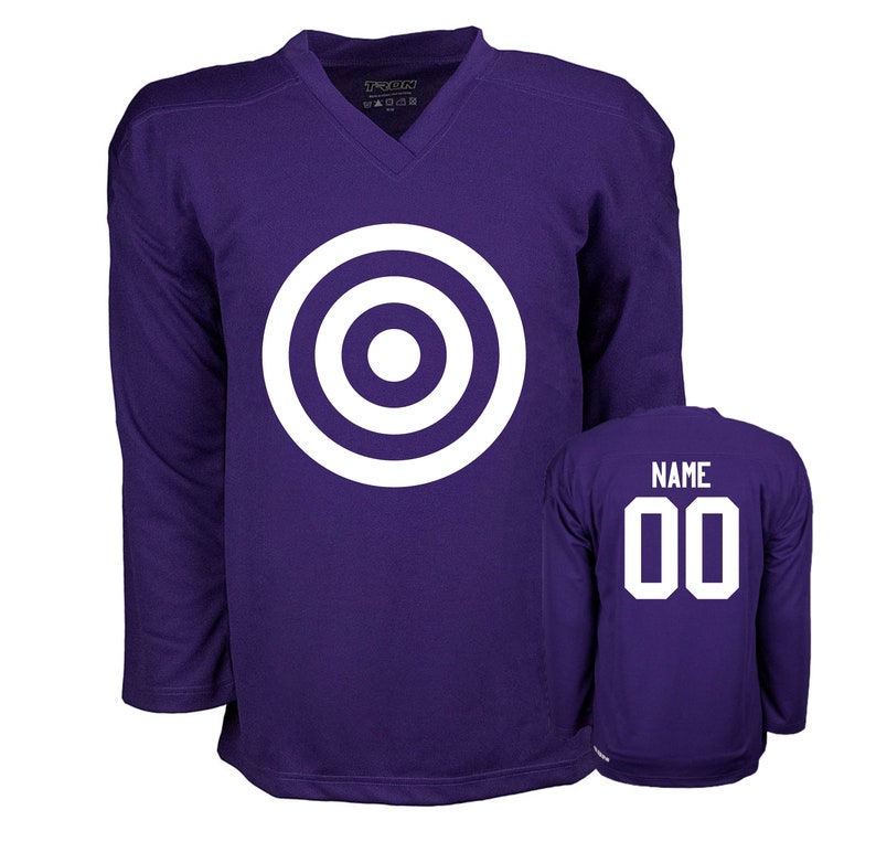 Puede incluir: Camiseta de hockey morada con un dise&ntilde;o de diana blanca en la parte delantera y el n&uacute;mero 00 en la parte trasera. La camiseta tiene cuello en V y mangas largas.