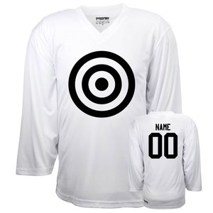 Puede incluir: Camiseta de hockey blanca con un dise&ntilde;o de diana en blanco y negro en la parte delantera y el n&uacute;mero 00 en la parte posterior. La camiseta tiene un cuello en V y mangas largas.