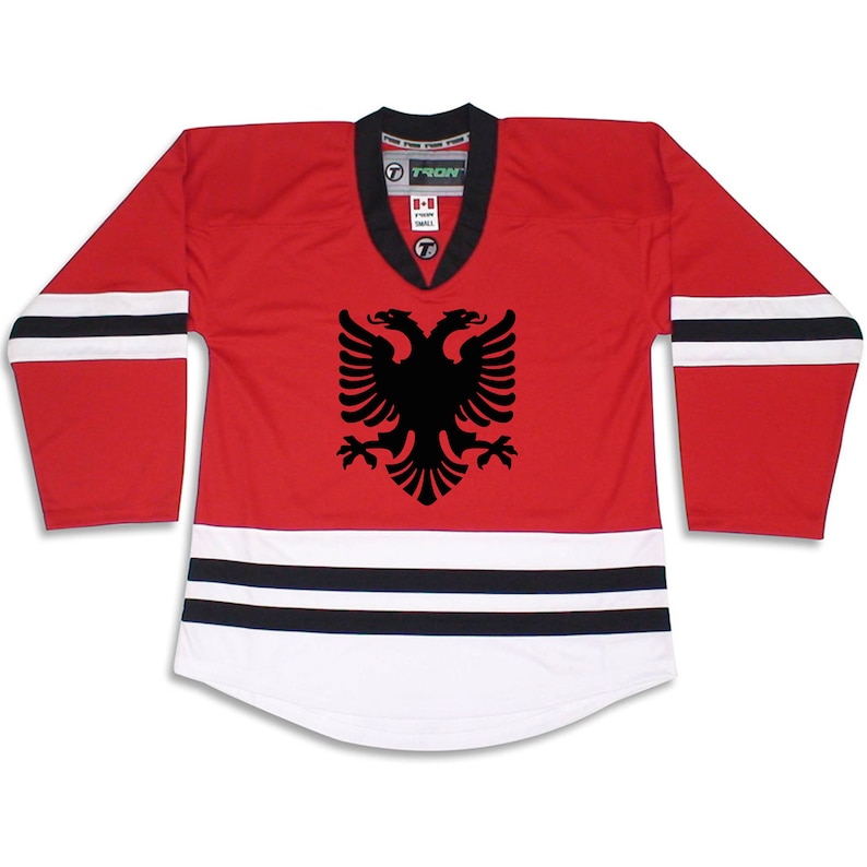 Puede incluir: Jersey de hockey rojo y blanco con un emblema de &aacute;guila bic&eacute;fala negra. El jersey tiene rayas negras y blancas en las mangas y en la parte inferior.