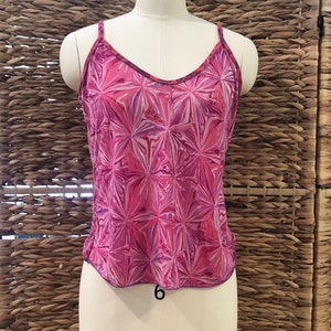 May include: A pink camisole with a V-neck and spaghetti straps. The top features a geometric pattern in shades of pink, purple, and white. The garment is displayed on a mannequin against a woven background.