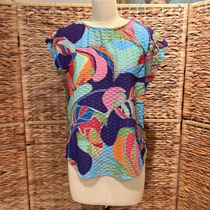Peut inclure: Un haut texturé et coloré avec un motif floral dans des tons de bleu, rose, orange et vert. Le chemisier à manches courtes est présenté sur un mannequin. Le haut a un col rond et un tissu texturé. Le fond est un écran tissé.