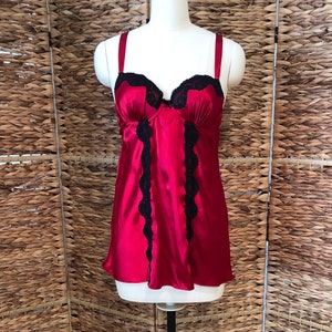 May include: A red satin camisole with black lace trim. The lingerie top features adjustable straps and a flattering silhouette. The black lace accents the neckline and the front of the garment. The camisole is displayed on a mannequin.