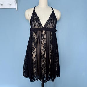 May include: Black lace lingerie dress on a mannequin. The dress features a V-neckline with delicate lace detailing and a flowing skirt. The dress is made of sheer fabric with floral lace accents.