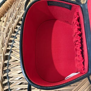 May include: Black oval-shaped handbag with a red interior. The bag has a black handle and a black trim. The interior has a red lining with a gathered edge. The bag is open, revealing the interior. The bag is sitting on a woven surface.