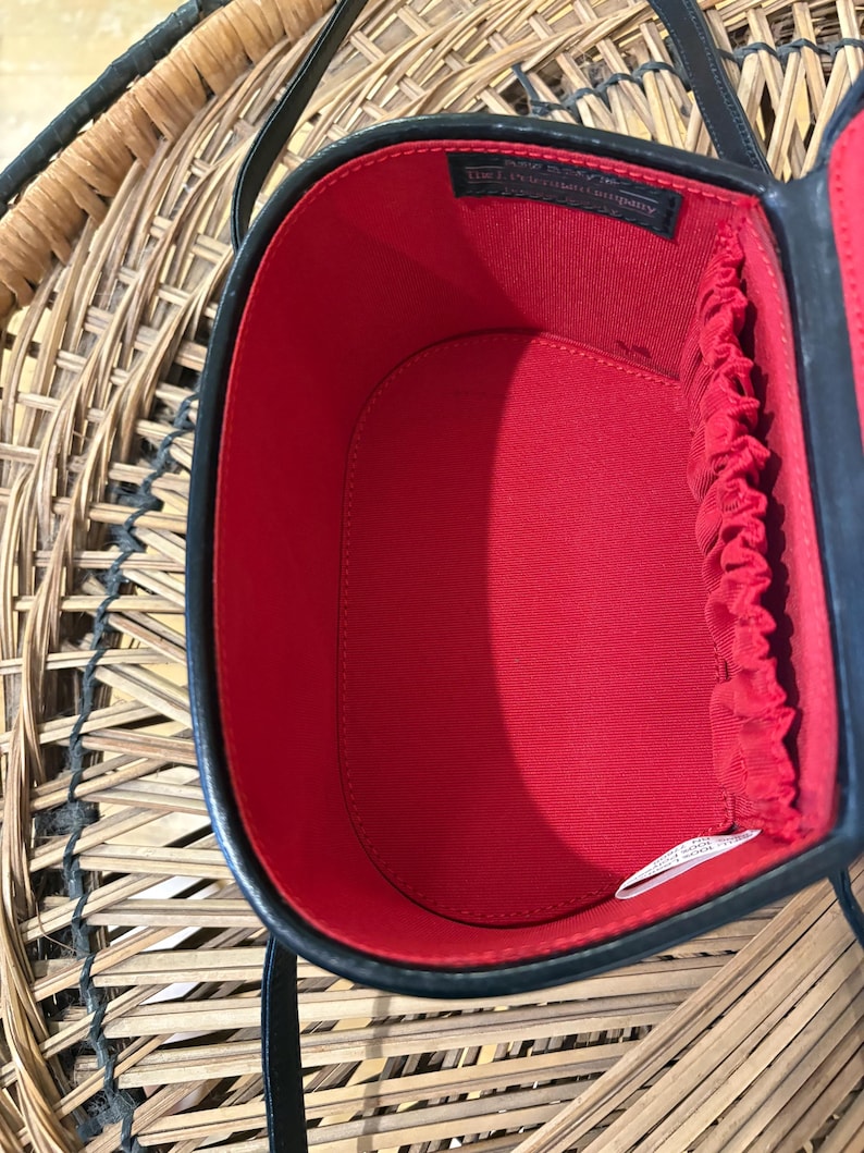 May include: Black oval-shaped handbag with a red interior. The bag has a black handle and a black trim. The interior has a red lining with a gathered edge. The bag is open, revealing the interior. The bag is sitting on a woven surface.