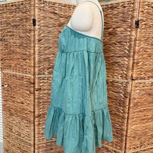 Op de afbeelding: Een turquoise mini-jurk met spaghettibandjes en een gelaagd ontwerp. De jurk wordt op een mannequin gepresenteerd, waarbij de textuur van de stof wordt benadrukt. Een lichte, zomerse kleding.