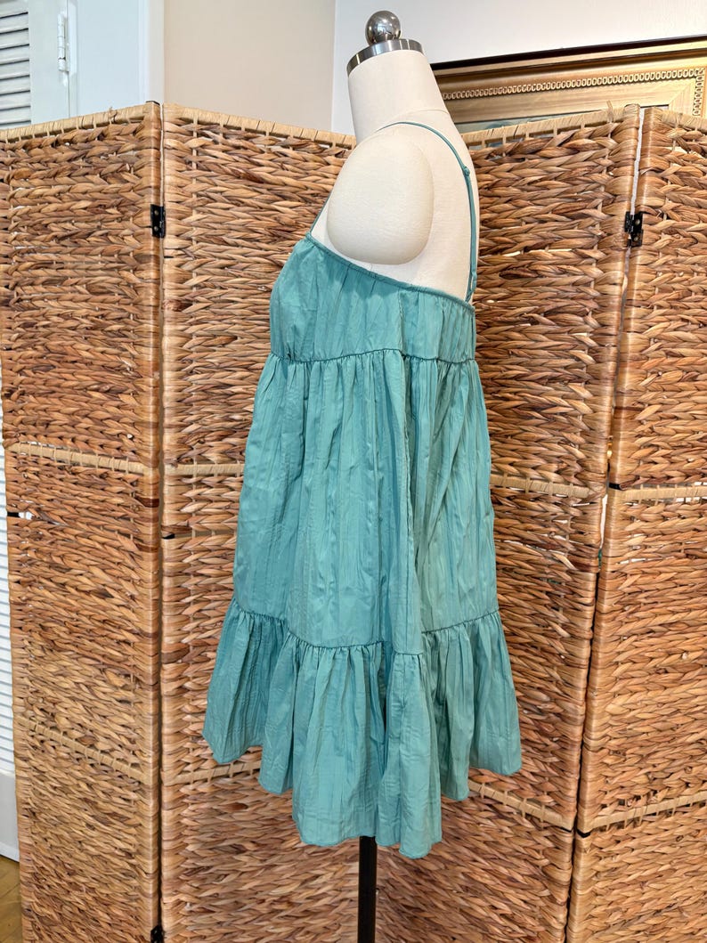 Op de afbeelding: Een turquoise mini-jurk met spaghettibandjes en een gelaagd ontwerp. De jurk wordt op een mannequin gepresenteerd, waarbij de textuur van de stof wordt benadrukt. Een lichte, zomerse kleding.