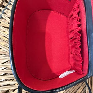 May include: Black oval-shaped handbag with a red interior. The bag has a structured design with a top handle and a shoulder strap. The interior features a small pocket and elastic bands. The bag is made of a glossy material.