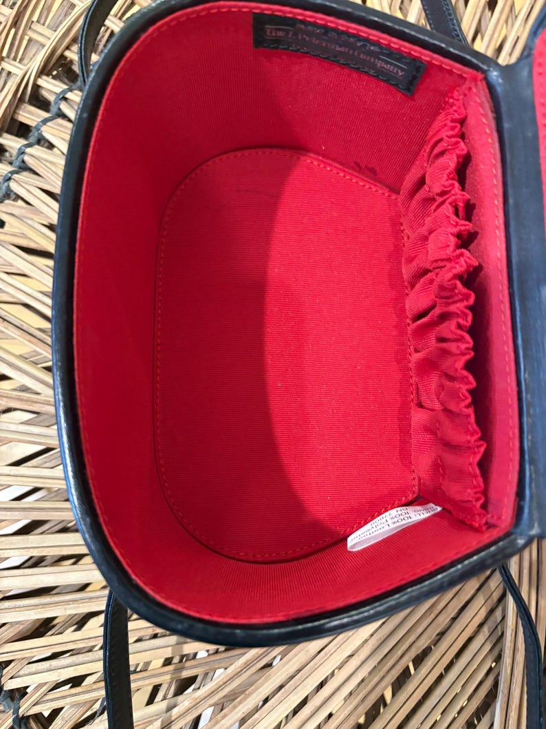 May include: Black oval-shaped handbag with a red interior. The bag has a structured design with a top handle and a shoulder strap. The interior features a small pocket and elastic bands. The bag is made of a glossy material.