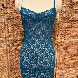 K&ouml;nnte beinhalten: Ein tealfarbenes Spitzenkleid mit Spaghettitr&auml;gern. Das Kleid hat ein florales Spitzendesign und einen gewellten Ausschnitt. Das Kleid wird auf einer Schaufensterpuppe vor neutralem Hintergrund gezeigt.