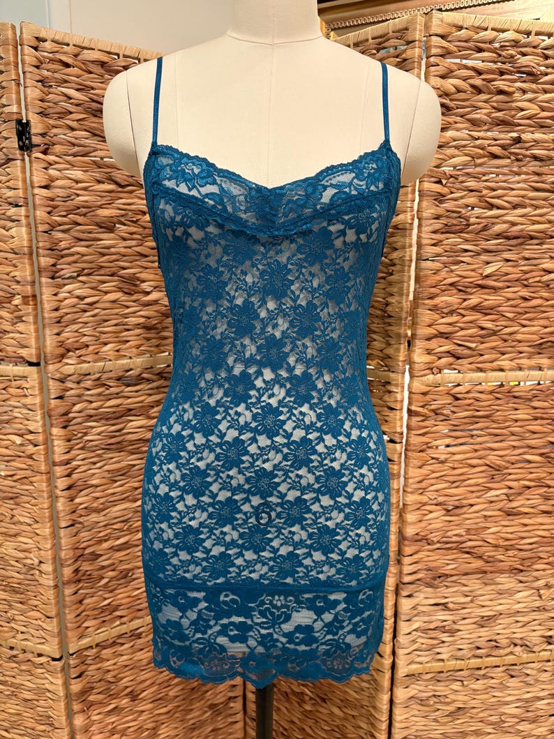 K&ouml;nnte beinhalten: Ein tealfarbenes Spitzenkleid mit Spaghettitr&auml;gern. Das Kleid hat ein florales Spitzendesign und einen gewellten Ausschnitt. Das Kleid wird auf einer Schaufensterpuppe vor neutralem Hintergrund gezeigt.