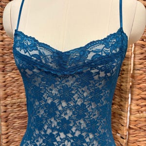K&ouml;nnte beinhalten: Ein tealfarbenes Spitzen-Camisole mit zarten Spaghettitr&auml;gern. Das Camisole hat ein florales Spitzenmuster und einen gewellten Ausschnitt. Das Kleidungsst&uuml;ck wird auf einer Schaufensterpuppe pr&auml;sentiert.